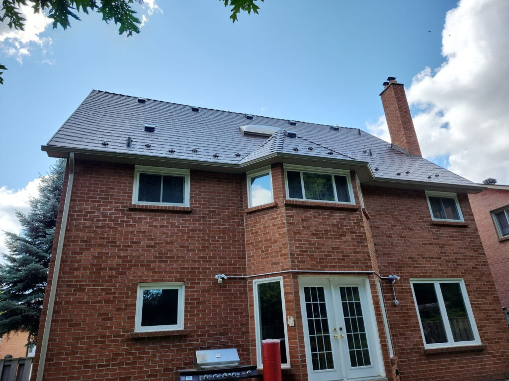 A two-story red brick house with white-framed windows, a grey metal roof, and a brick chimney, viewed from the backyard under a partly cloudy sky.