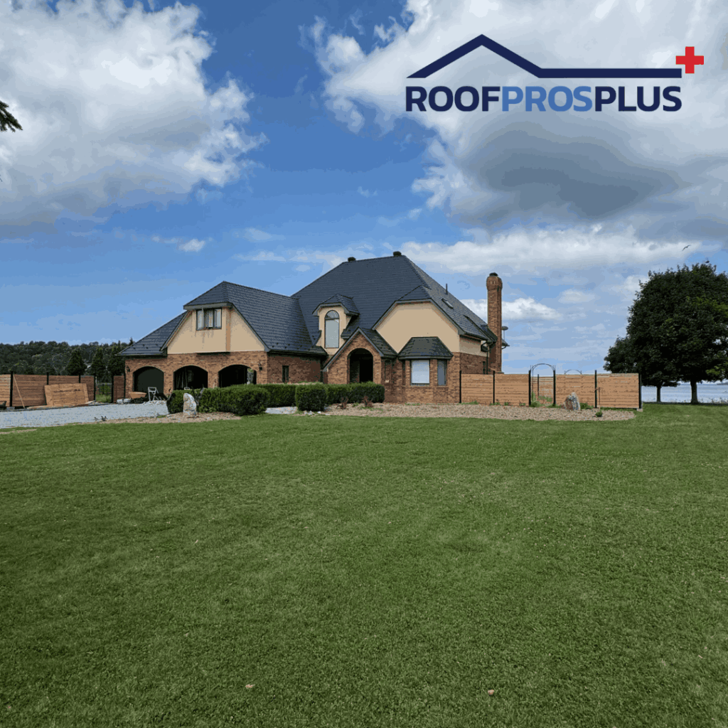 A large brick house with dark metal roofing in a spacious green yard. There is a blue sky backdrop with scattered clouds. The "RoofProsPlus" logo is in the top right corner.