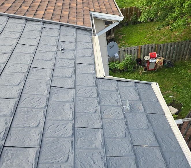 A grey metal rooftop is adjacent to a brown asphalt shingle roof. Below, a fenced green yard has a satellite dish and outdoor items in it.