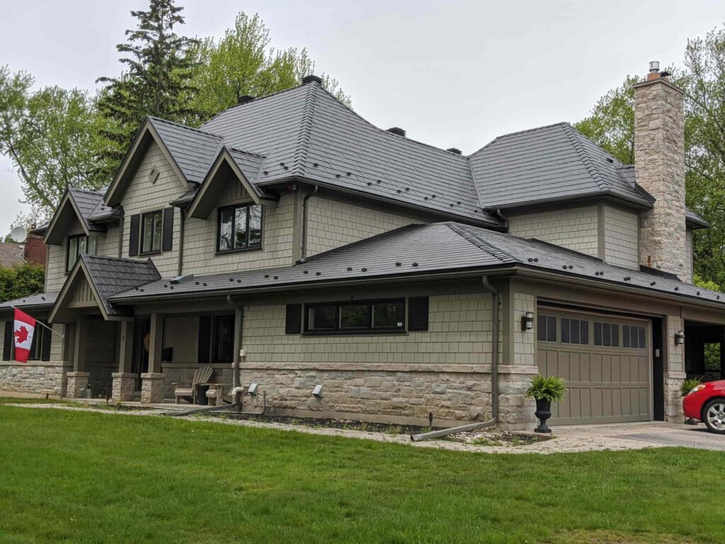 A two-story house with a stone and metal exterior featuring a modern gray roof and a prominent stone chimney. The property includes a garage, a neatly maintained lawn, a Canadian flag on display, and a bright red car partially visible in the driveway.