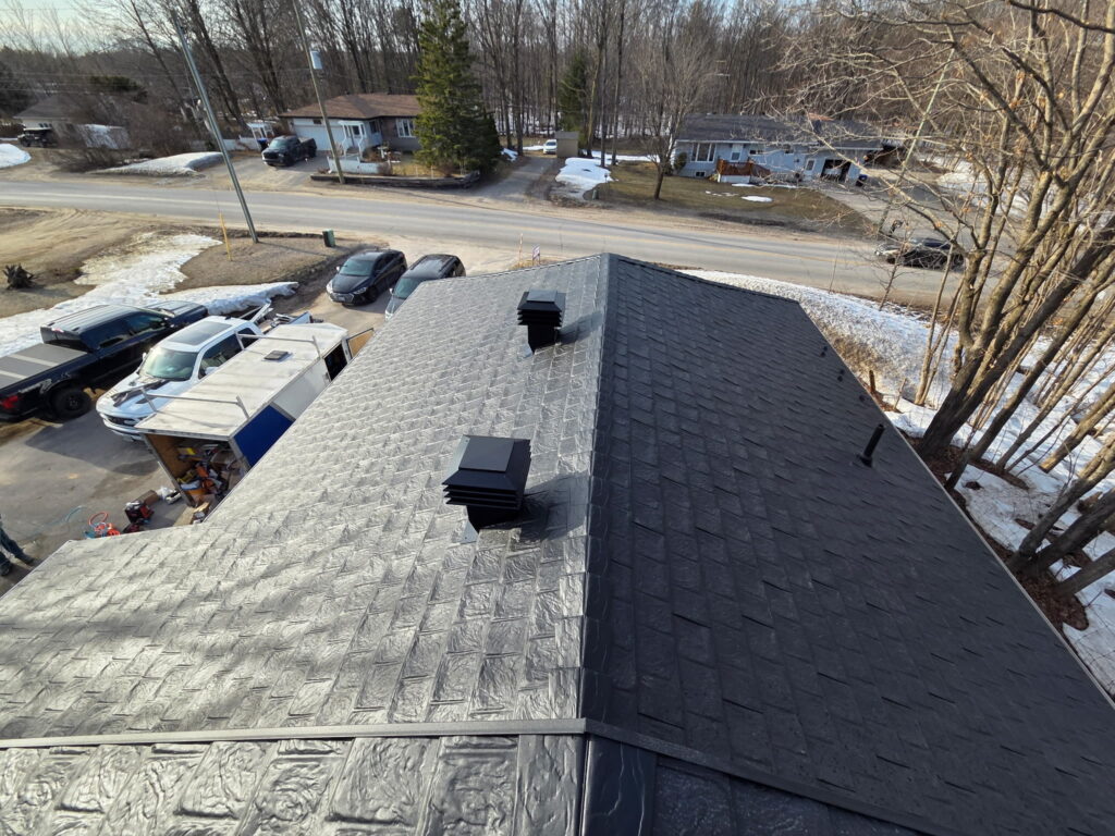 Aerial view of a house's dark metal roof with two vents, surrounded by snowy yards and parked cars. Trees line a clear, lightly snow-covered road.