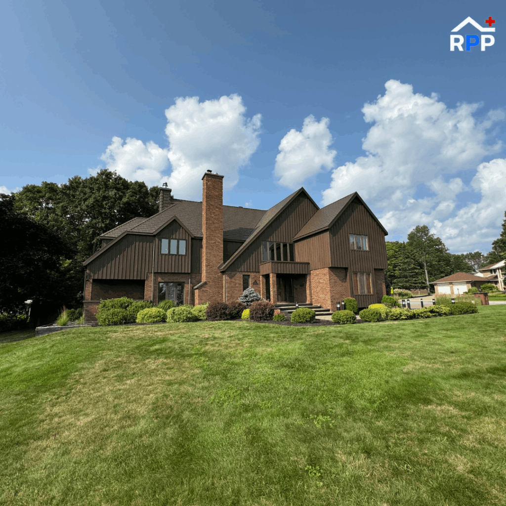 A large brown brick house with dark wood accents and a metal roof, is surrounded by lush green lawn and shrubs. A blue sky with fluffy clouds adds a serene backdrop.