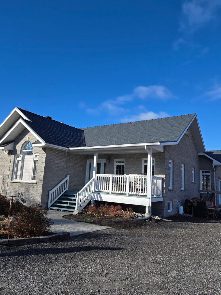 One-story brick house with a large porch, white railings, and a dark metal roof sits under a clear blue sky. Landscaping with brown shrubs is in front of the house.