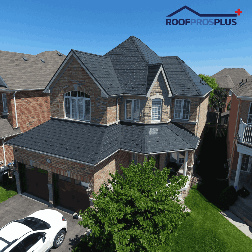 A two-story brick house with a dark, textured metal roof under a clear blue sky. The roof has various angles and gables. A white car is parked in the driveway.