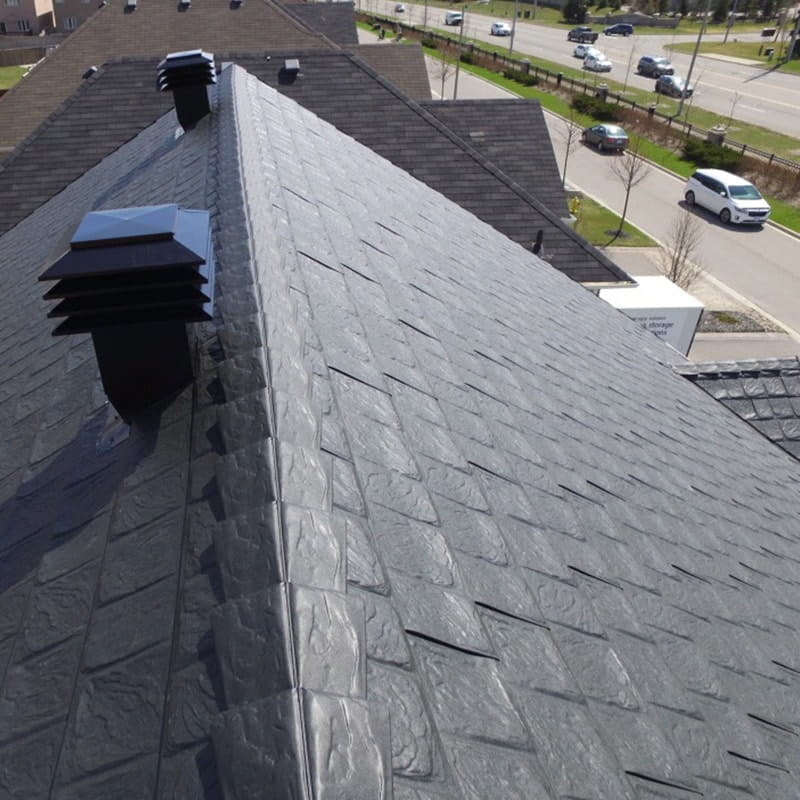 Close-up picture of a textured, grey metal roof with black vents, overlooking a suburban street with cars and greenery. 