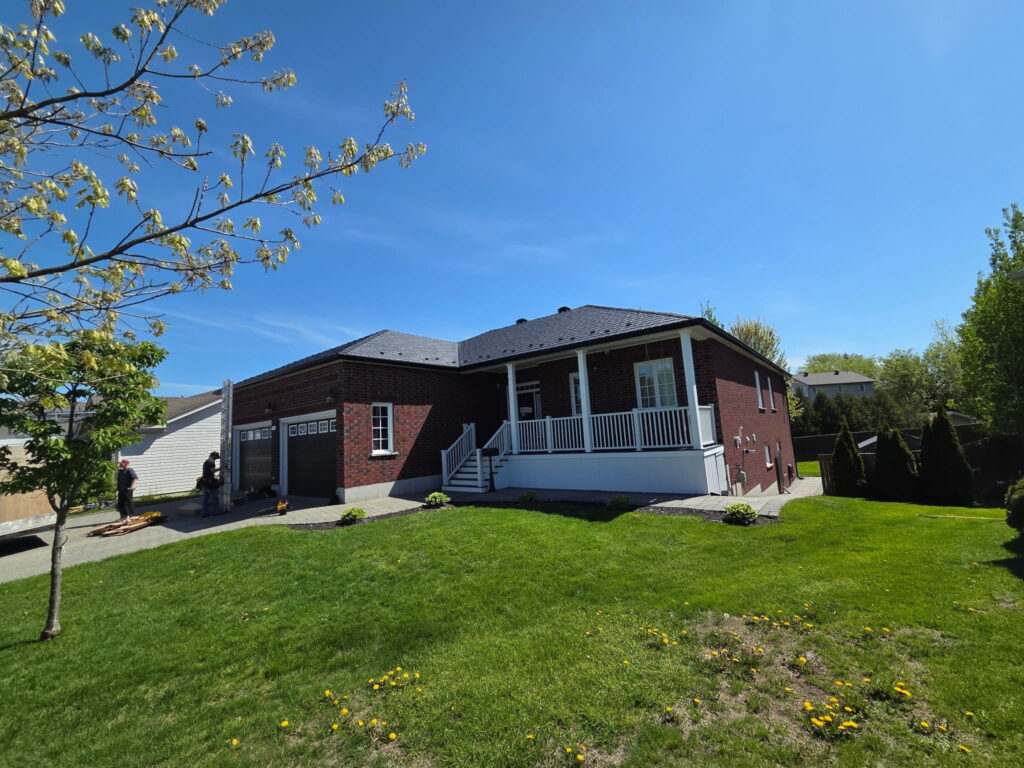 A brick house with a front porch, white railing, and a two-car garage is set against a clear blue sky. A well-kept lawn with a tree is standing in the scene.