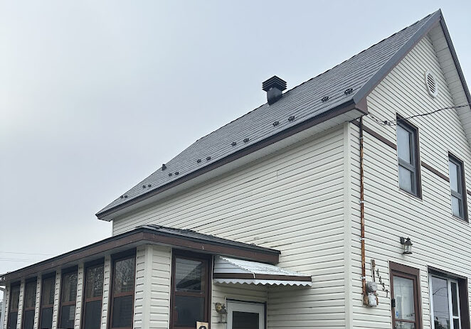 A two-story house with beige siding, a dark metal roof, and a small enclosed porch.