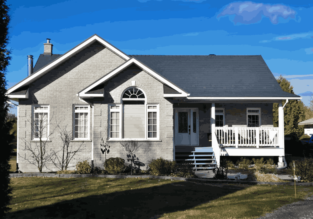 A single-story grey brick house under a clear blue sky, with a dark metal roof. The house has white trim, arched windows, and a small front porch.