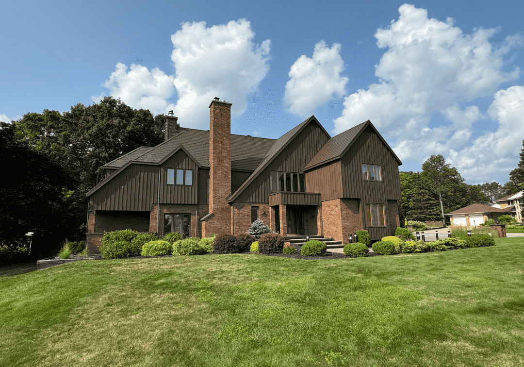 A large brick home with dark metal roofing in Ontario, showcasing Canadian-made roofing built for durability and style against a bright blue sky with clouds. The "Roof Pros Plus" logo is in the top right corner of the image.