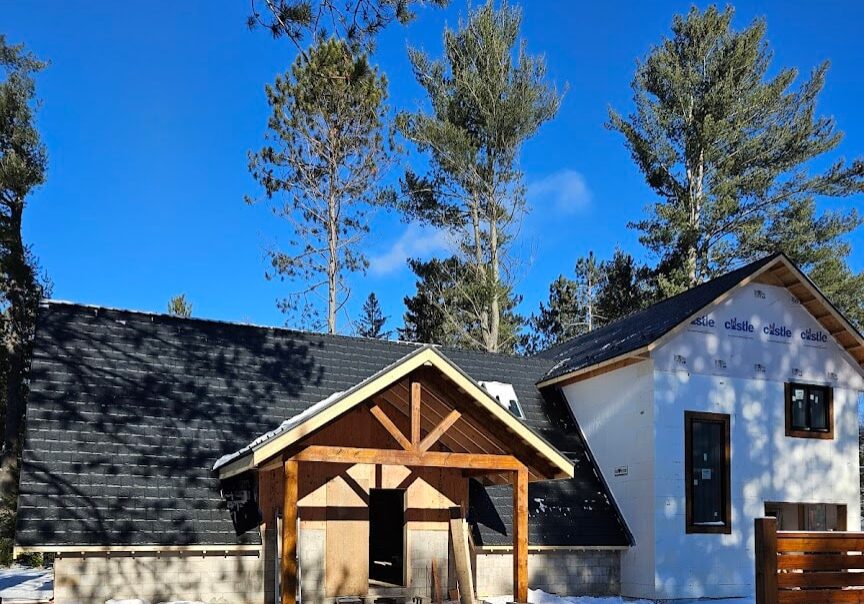 A house is enclosed by a wooden fence, with a wooden gate providing access to the property. A dark, sloped metal roof is the feature point of the house.
