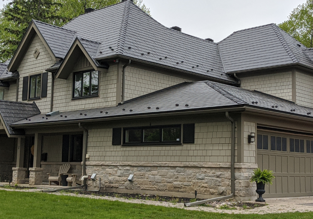 A beautiful house with a grey metal roof, stone and siding exterior, attached garage, and the Roof Pros Plus logo in the corner, which is an expert in metal roof installation and quality roofing materials.