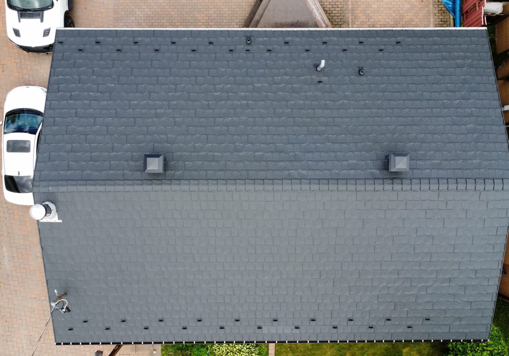 Aerial view of a grey metal roof with two small chimneys. Two white cars are parked on a brick driveway beside landscaped greenery.