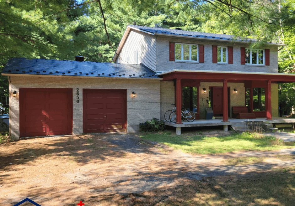 A two-story house with a newly installed metal roof, light brick exterior and red trim, surrounded by lush trees. A porch with bicycles and a shaded driveway are at the front of the house.