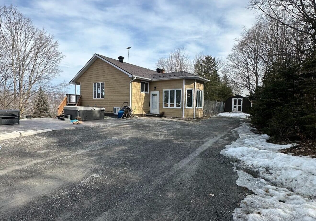 Small beige house with a metal roof and a porch in a winter setting. Snow lines the gravel driveway. Bare trees surround the house under a partly cloudy sky.