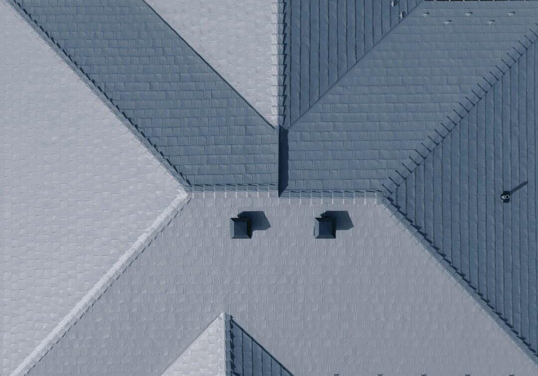 Aerial view of a grey metal roof with two vents, a white car parked in the driveway, and a logo that reads "ROOF PROS PLUS Inc." at the top of the image.