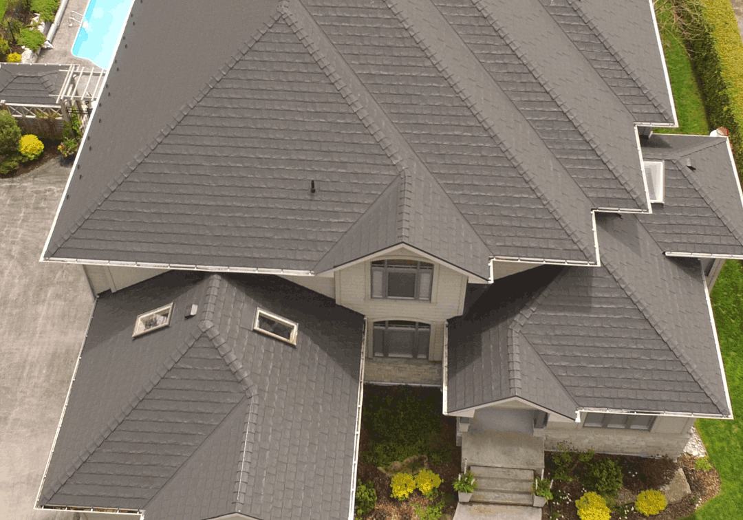 Aerial view of a large house with a dark gray, multi-gabled metal roof. A swimming pool and green lawn are visible in the backyard.