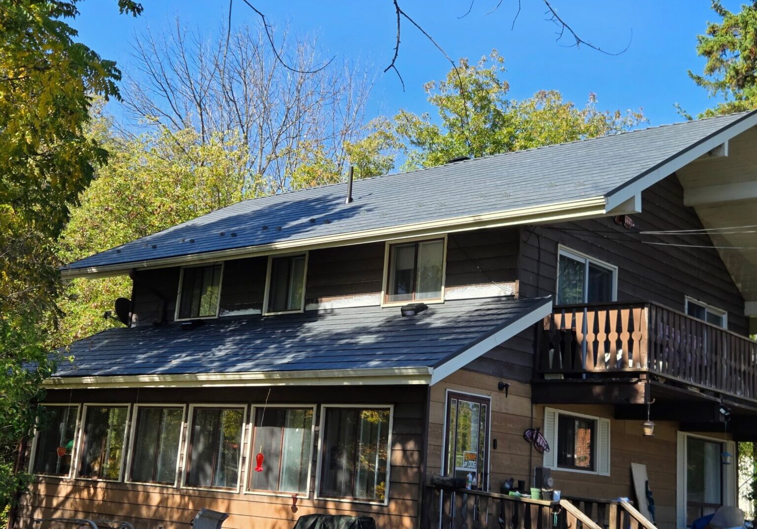 A two-story brown wooden house with a blue-grey metal roof, surrounded by lush trees under a clear blue sky.