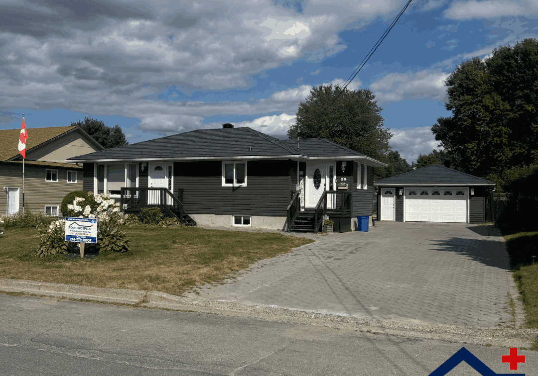 Single-story brown house with black metal roof, driveway, and detached garage. Canadian flag, for-sale sign, and flower bushes are in the front yard under a partly cloudy sky.