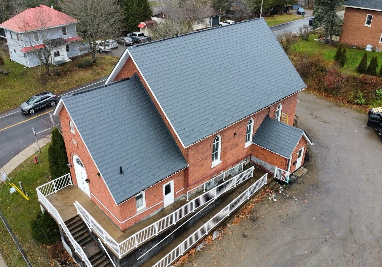 Aerial view of a brick building with a steep dark metal roof, adjacent white railings, and surrounded by autumn trees.