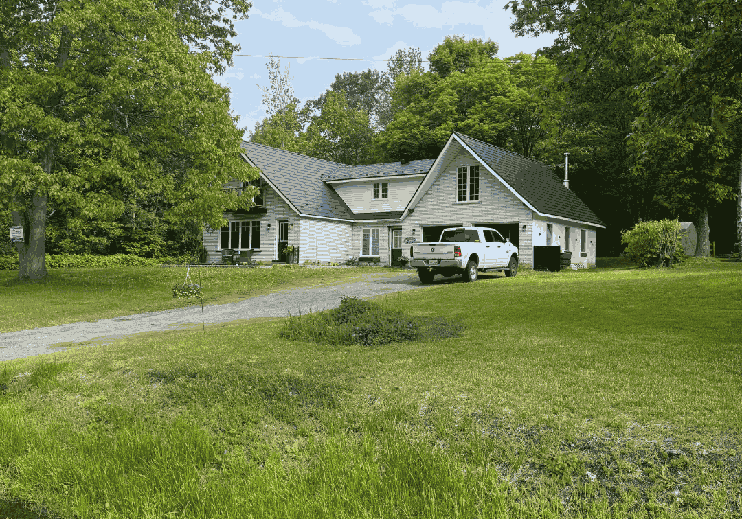 A white house with a dark metal roof is surrounded by trees and greenery. A white truck is parked on the gravel driveway. The scene is peaceful and rural. Logo text reads "ROOFPROSPLUS" at the top.