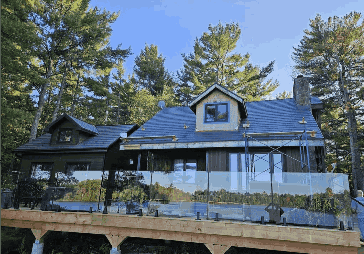 A rustic cabin with a dark roof under construction, surrounded by tall pine trees. It features a wooden deck with glass railings, set against a clear blue sky.