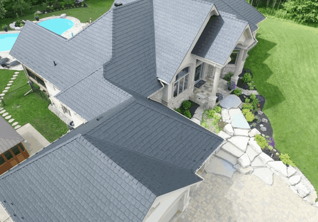 Aerial view of a modern house with a dark grey metal roof, surrounded by lush green lawn and trees. A pool is visible in the backyard.