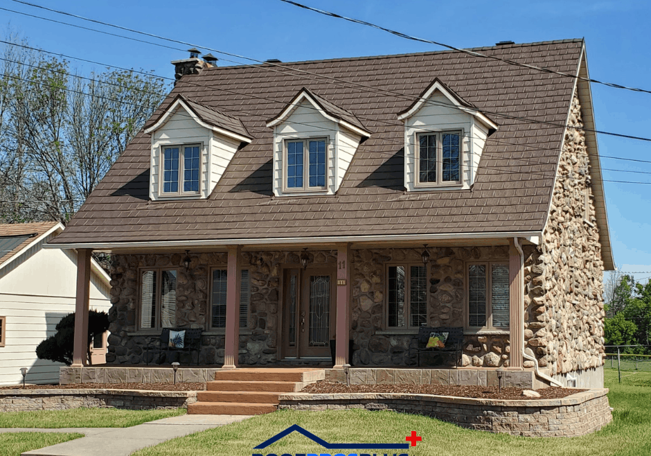 A stone house with a brown metal roof and three dormer windows, surrounded by green lawn. A porch with columns and steps leads to the entrance.