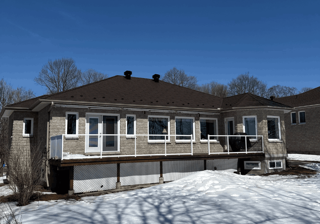 A modern house with a brown metal roof sits against a clear blue sky. The snow-covered yard creates a crisp contrast. The RoofProsPlus Inc.logo is at the top of the image.