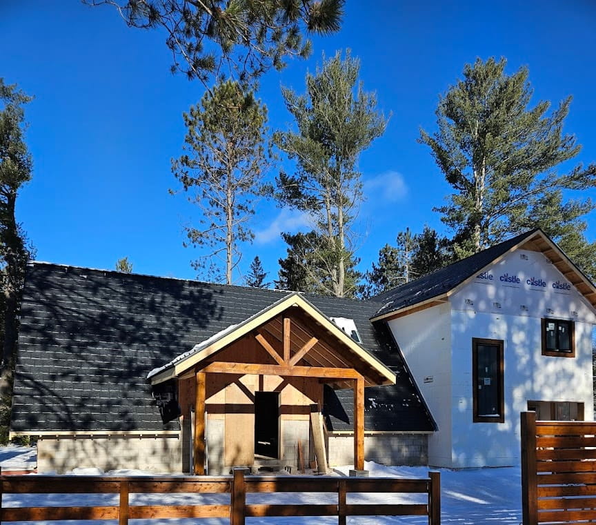 A partially constructed house with a black metal roof and wooden entrance stands in a snowy landscape. Tall pine trees and a bright blue sky frame the scene.