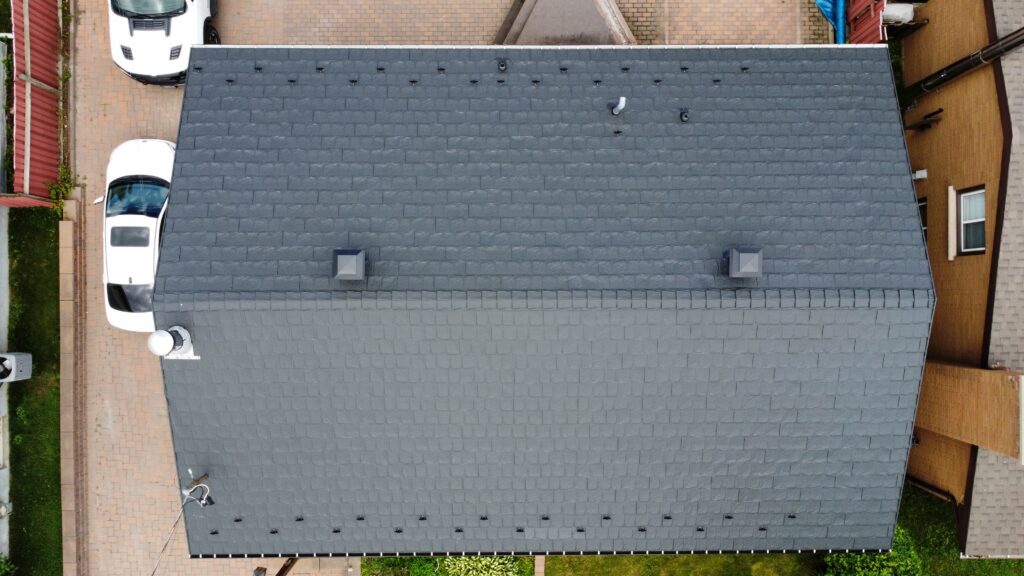 Aerial view of a grey metal roof with two small chimneys. Two white cars are parked on a brick driveway beside landscaped greenery.