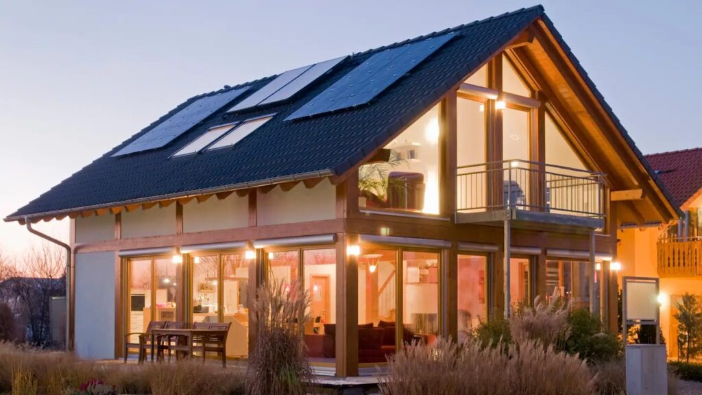 A beautiful residential home with many large windows and solar panels on the roof. The picture is taken during dusk, and the interior of the house is lit up from lights.