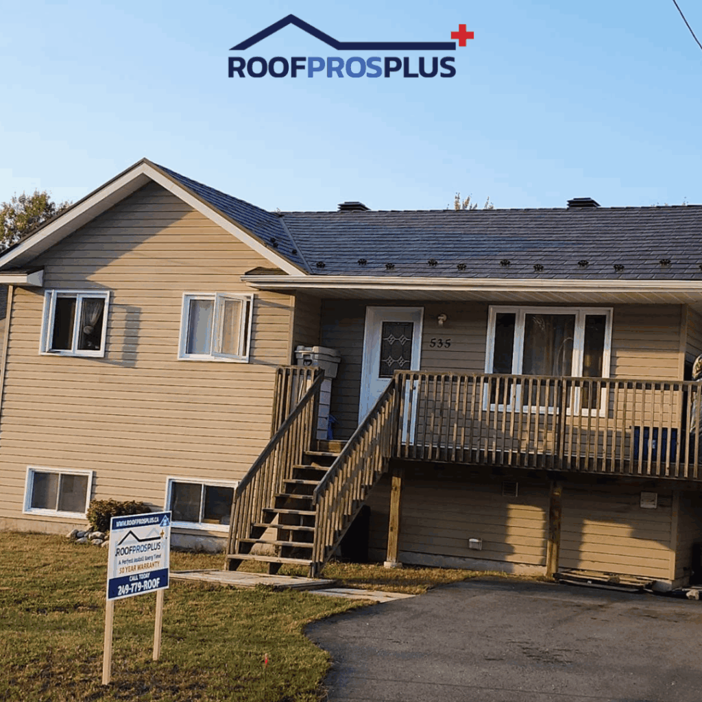A two-story beige house with a newly shingled metal roof and front porch stairs. A "RoofProsPlus" yard sign stands on the grassy lawn under a clear sky.