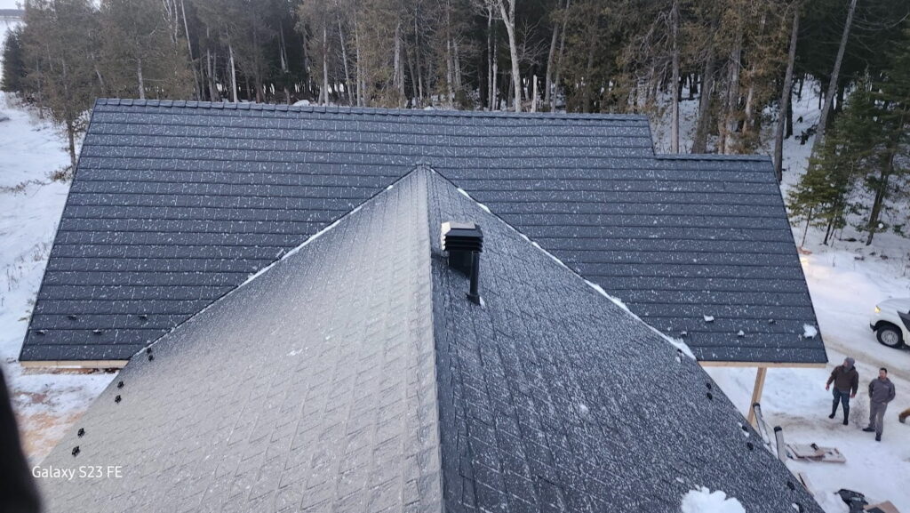 A snow-dusted, newly installed metal roof of a house surrounded by a snowy forest, with two people and a vehicle in the bottom right corner. 