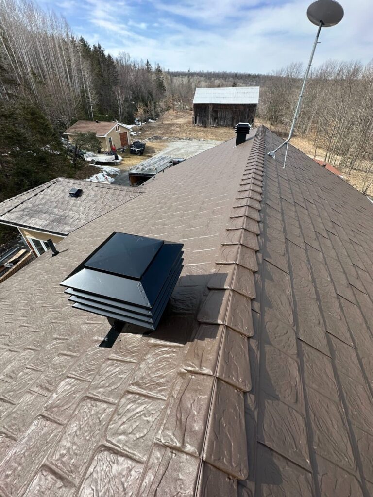 Brown metal roof with a vent and a satellite dish, viewed along the ridge. Surrounding landscape includes trees, a barn, and outbuildings under a blue sky.