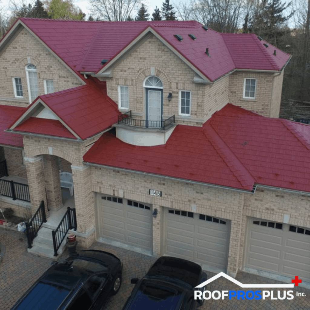 A two-story brick house with vibrant red metal roofing, arched windows, and a small balcony. The garage doors are beneath the balcony. Trees and a cloudy sky are in the background.