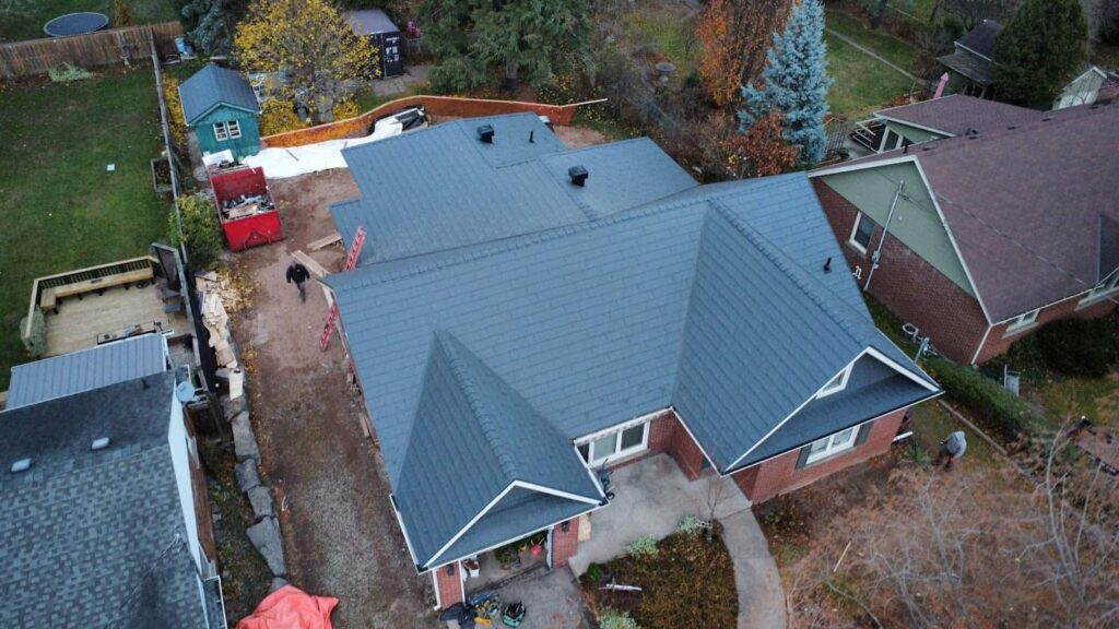 Aerial view of a red brick house with a grey metal roof, surrounded by a yard with trees in autumn colors. A person works by the side of the house, among construction materials.