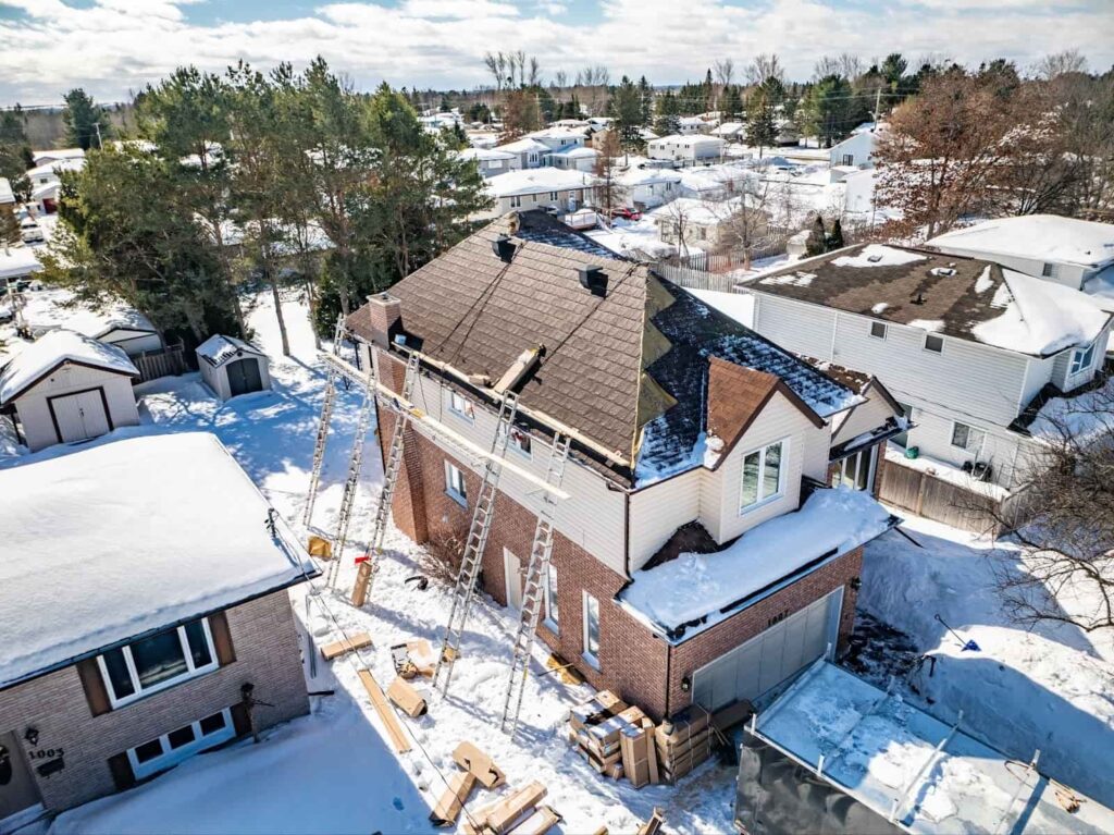 Aerial view of a snow-covered suburban house undergoing metal roof repairs, with ladders propped against the sides and construction materials on the ground.