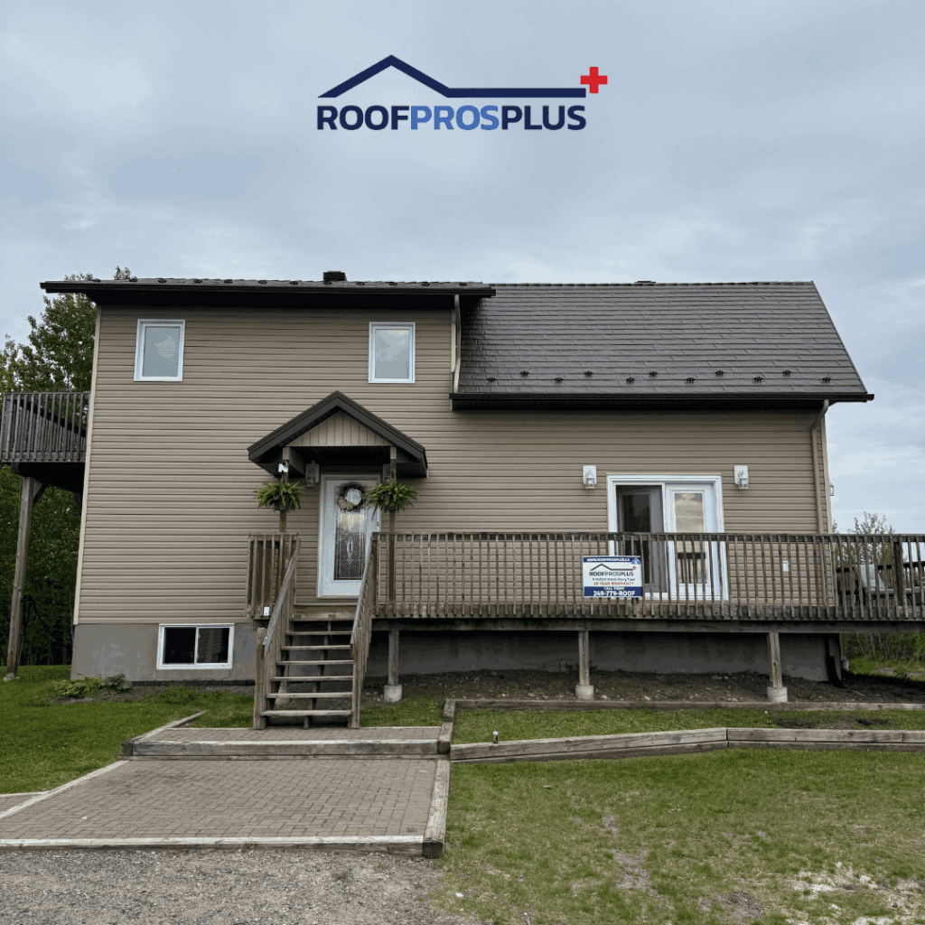 A two-story house with beige siding and a dark metal roof under a cloudy sky. A sign on the porch reads "RoofProsPlus."