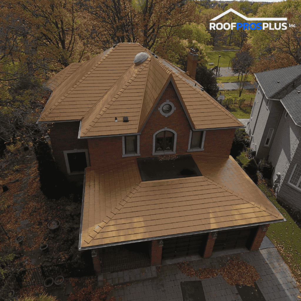Aerial view of a two-story house with a brown sloped metal roof. Surrounding trees show autumn foliage, and a driveway leads to a garage at the front. The house features multiple windows and a decorative facade.