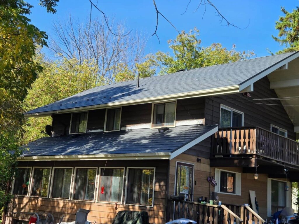 A two-story wooden house with dark siding and a sloped roof is surrounded by trees under a clear blue sky. Sunlight casts gentle shadows, adding warmth.