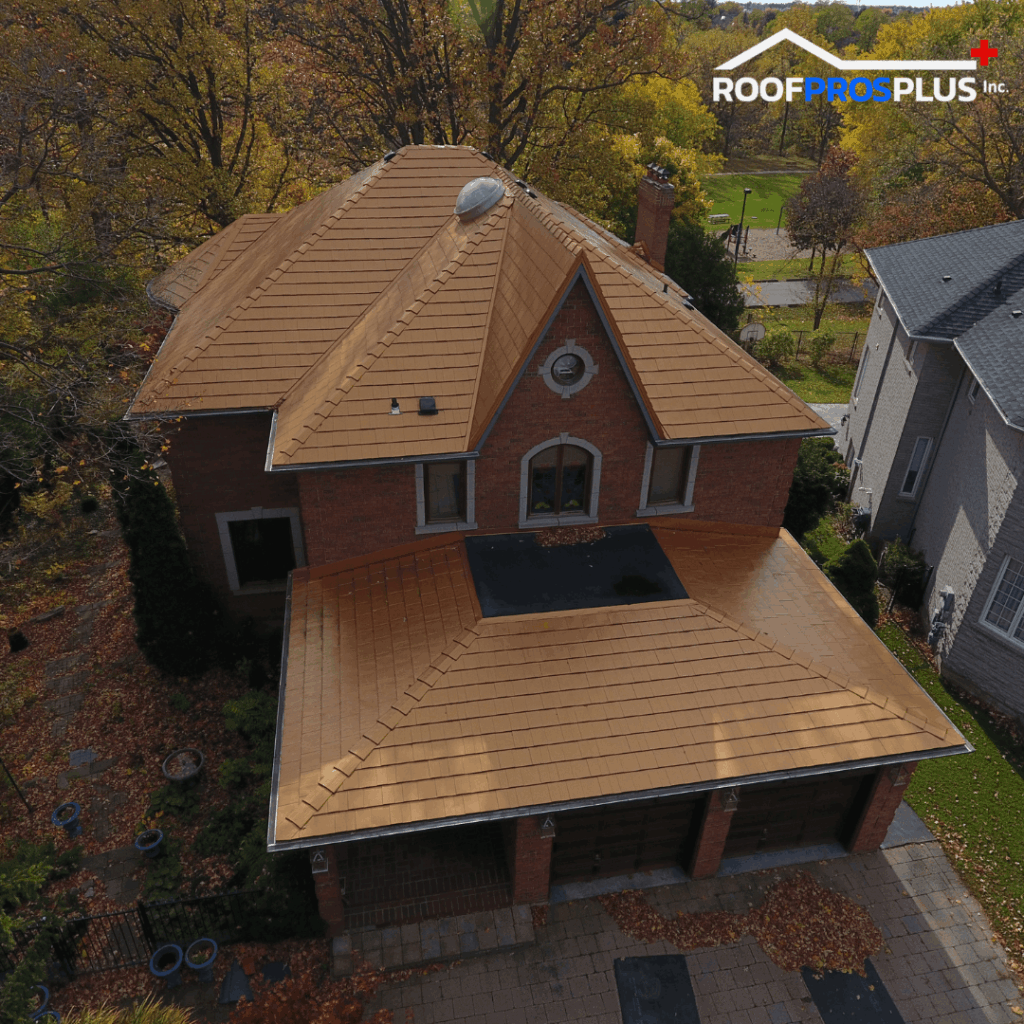 Aerial view of a two-story brick house with a new brown metal roof surrounded by autumn trees. The logo "ROOF PROS PLUS" is on the top right. Leaves scatter the driveway.