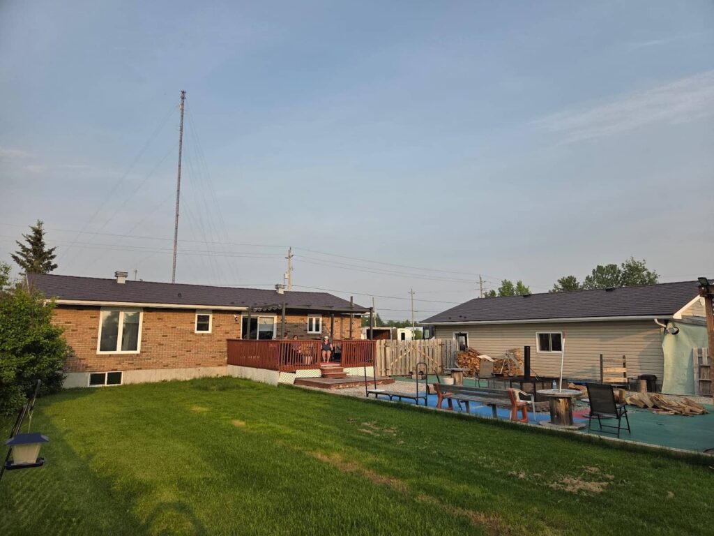 A backyard scene with a grassy lawn, two brick and siding houses, a wooden deck, and an above-ground pool. 