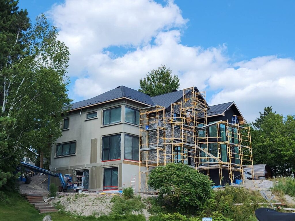 A large house under construction is surrounded by scaffolding. It sits amid lush greenery under a blue sky with fluffy clouds.