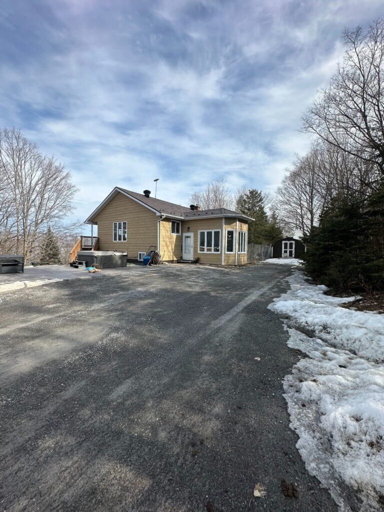 Small beige house with a metal roof and a porch in a winter setting. Snow lines the gravel driveway. Bare trees surround the house under a partly cloudy sky.