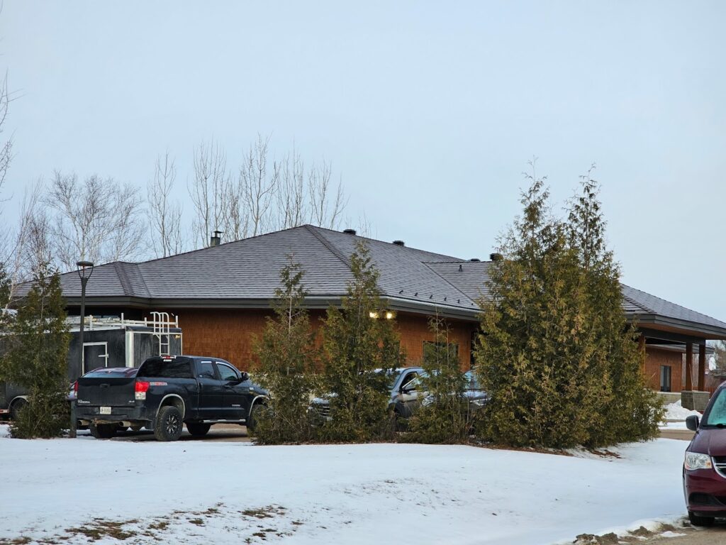 A single-story brown building with a grey metal roof, surrounded by evergreen trees. Parked cars on a snowy ground suggest a cold, overcast winter day.