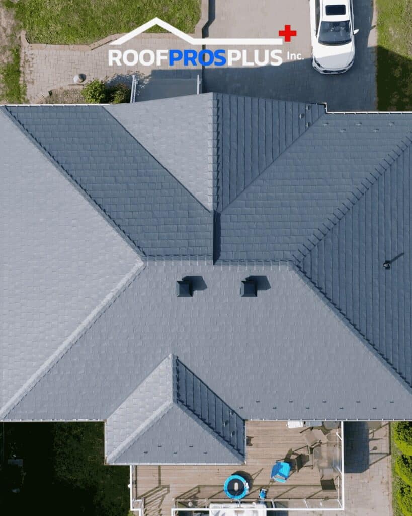 Aerial view of a grey metal roof with two vents, a white car parked in the driveway, and a logo that reads "ROOF PROS PLUS Inc." at the top of the image.