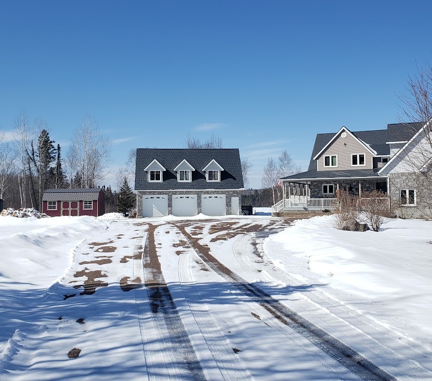 A snowy driveway leads to a large grey house with a three-car garage and a metal roof. A red shed stands to the left, surrounded by bare trees under a clear blue sky.