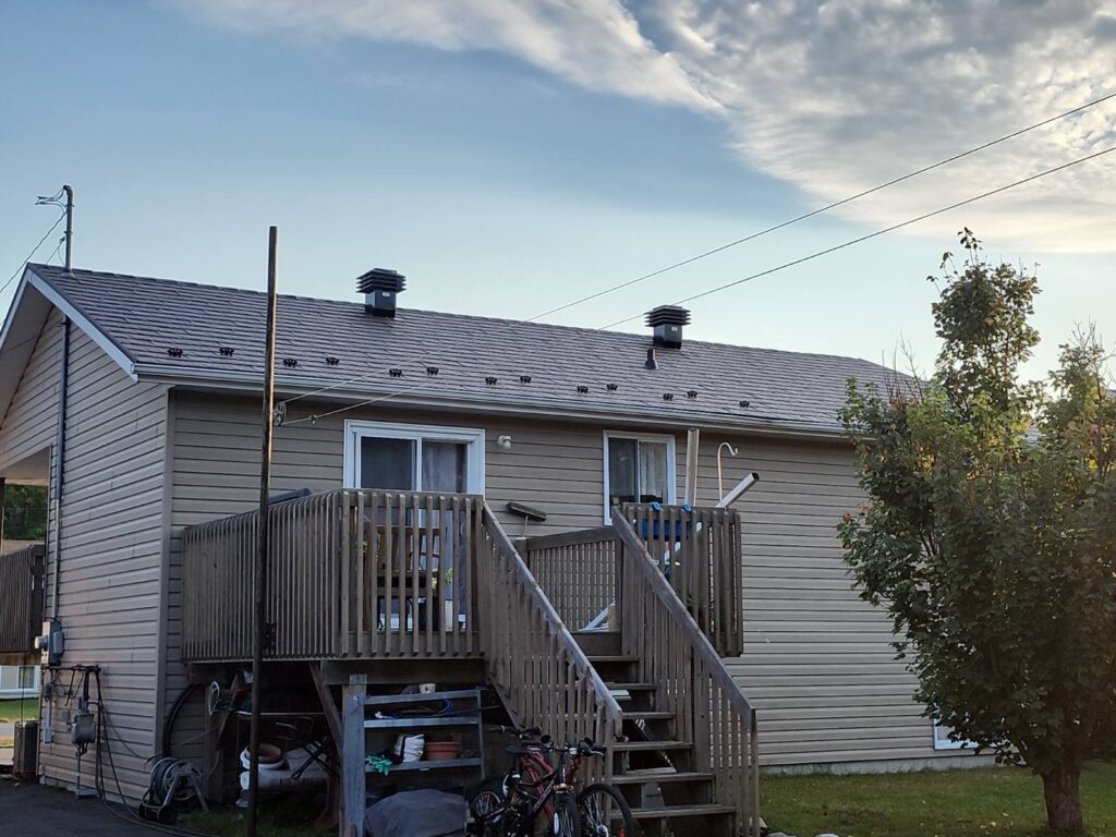 A single-story house with beige siding, a wooden deck, and stairs. Bikes and items are stored underneath. A tree and a cloudy sky are in the background.