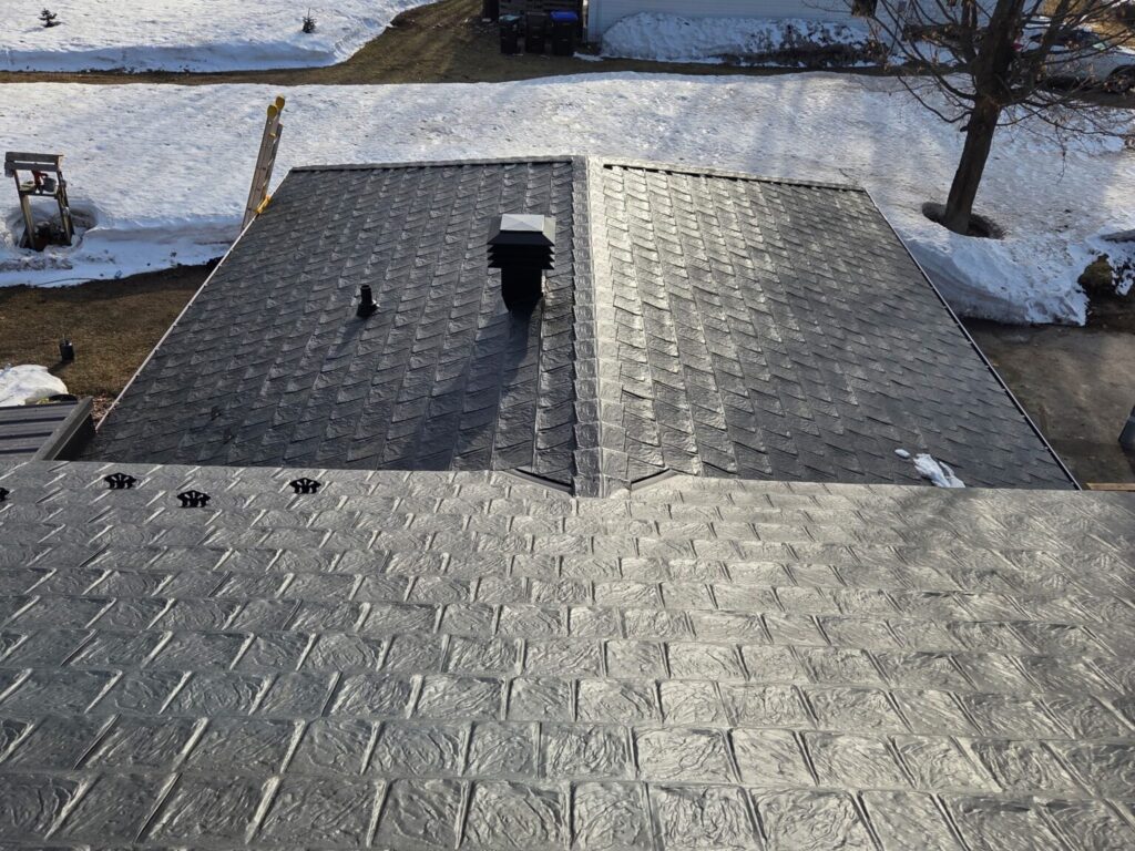 Aerial view of a textured, dark metal roof with snow-covered ground surrounding it. A chimney and two vents are visible. The scene is calm and wintery.