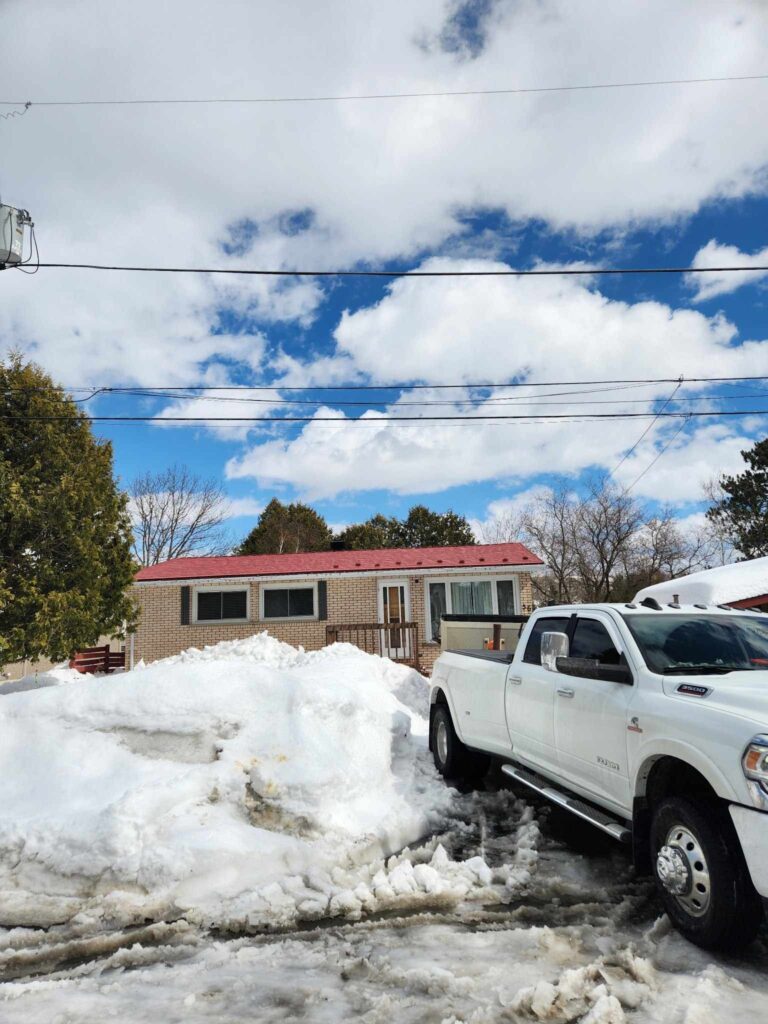 A snowy scene with a white pickup truck parked in front of a small house with a red metal roof. The sky is bright blue with fluffy clouds.
